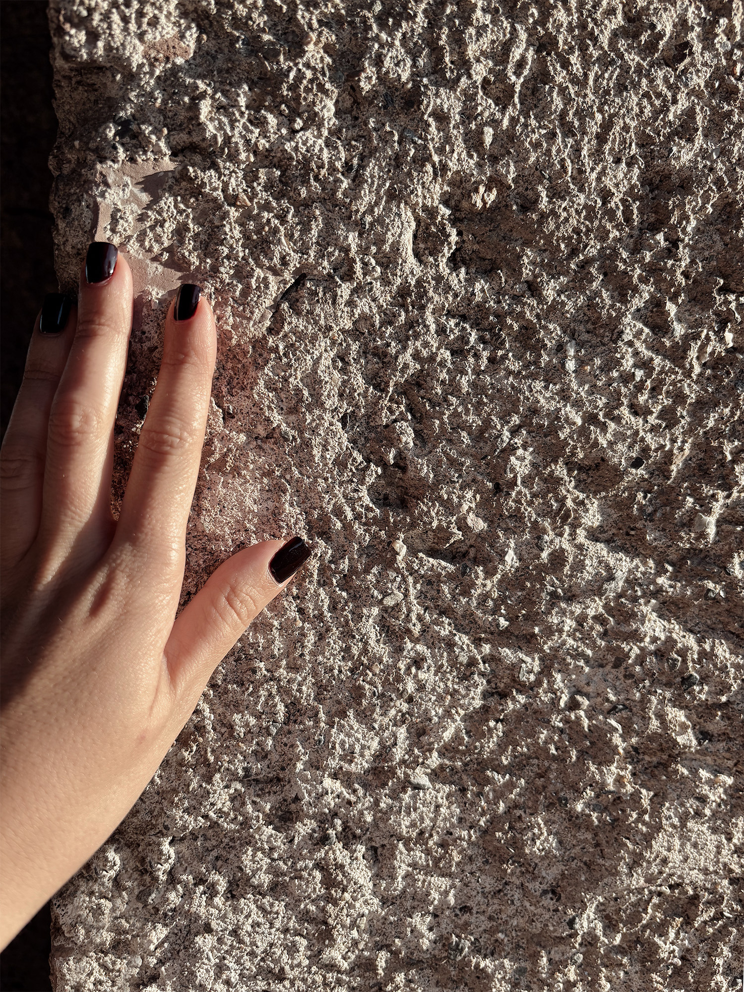 A hand resting against a rough, weathered wall in low sunlight, its coarse surface catching light and shadow